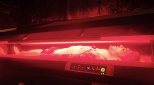 MMA fighter Rami Hamed lies in a NovoTHOR red light therapy pod at Acupuncture Zen, bathed in a soft red glow. He’s wearing athletic shorts, with his eyes closed, appearing relaxed and focused. The futuristic pod, designed for whole-body photobiomodulation, surrounds him, promoting recovery and wellness as he prepares for BRAVE CF 100. The setting is clinical yet serene, with dim lighting highlighting the pod’s sleek design.