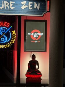 Exterior entrance of Acupuncture Zen clinic in Delray Beach, featuring illuminated "ACUPUNCTURE ZEN" sign, neon "NeedleYou.com" display, and a glowing red-lit Buddha statue under a wooden signboard.