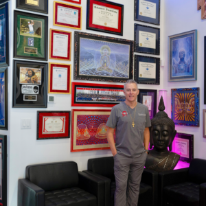 Dr. Tony Willcox standing proudly in front of his wall of credentials and autographed memorabilia at Acupuncture Zen clinic in Delray Beach, Florida, including university degrees, certifications, PGA and UFC endorsements, and spiritual artwork with a large Buddha statue nearby.
