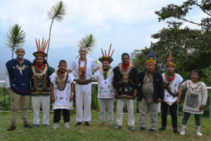 Dr Tony Willcox standing with a group of Taitas from the Inga lineage at Finca Ambiwasi, Colombia. The healers wear traditional ceremonial dress including feathered headdresses and bone necklaces, with mountain landscape in the background.
