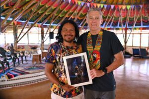 Dr Tony Willcox and Taita Juanito standing together at Finca Ambiwasi in Colombia, inside a colourfully decorated ceremonial space. Taita Juanito holds a portrait painting, and Dr Tony wears a traditional beaded ceremonial sash.