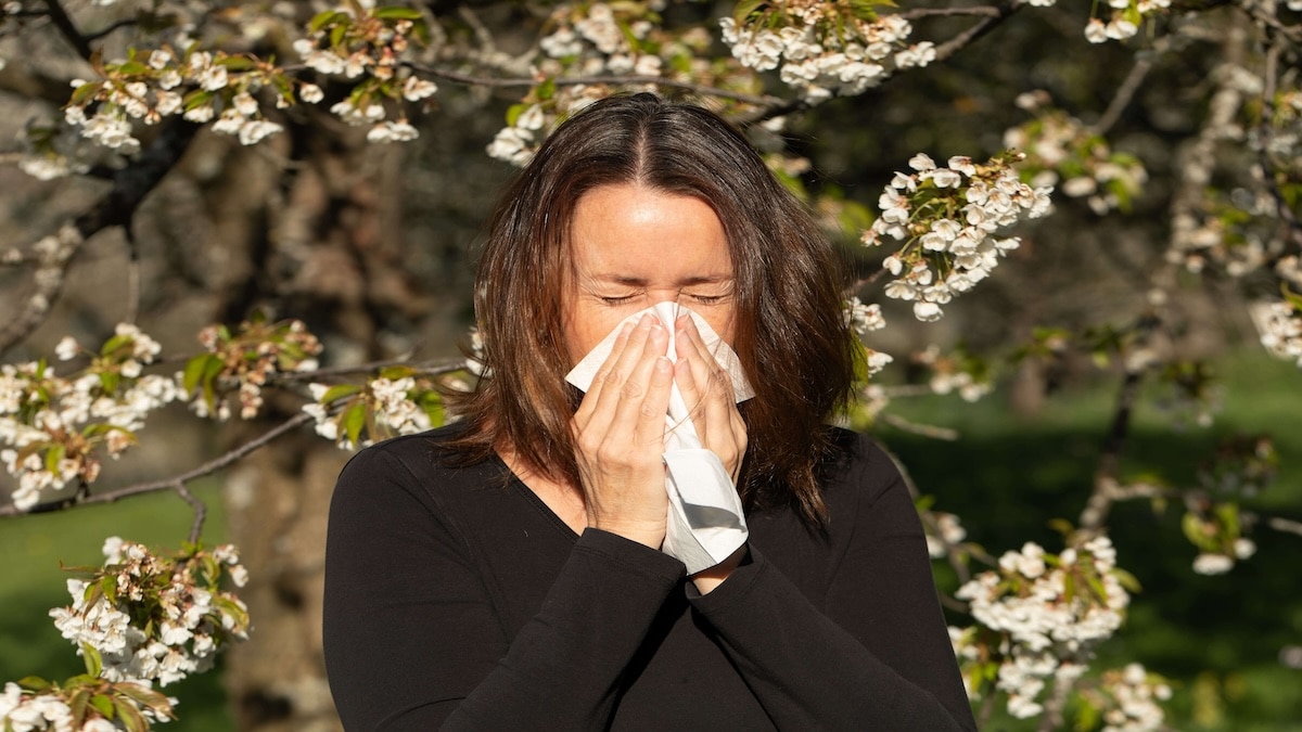 Woman blowing her nose into a tissue outdoors beneath blossoming spring trees, experiencing allergic rhinitis symptoms