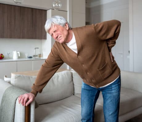Older man holding his lower back in pain at home, a common experience with lumbar spinal stenosis