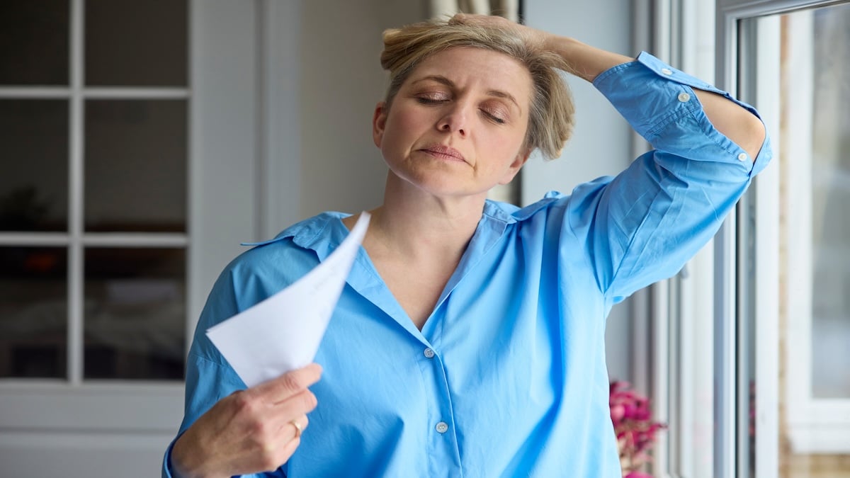 Middle-aged woman fanning herself with paper during a hot flash by a window, menopause acupuncture in Delray Beach